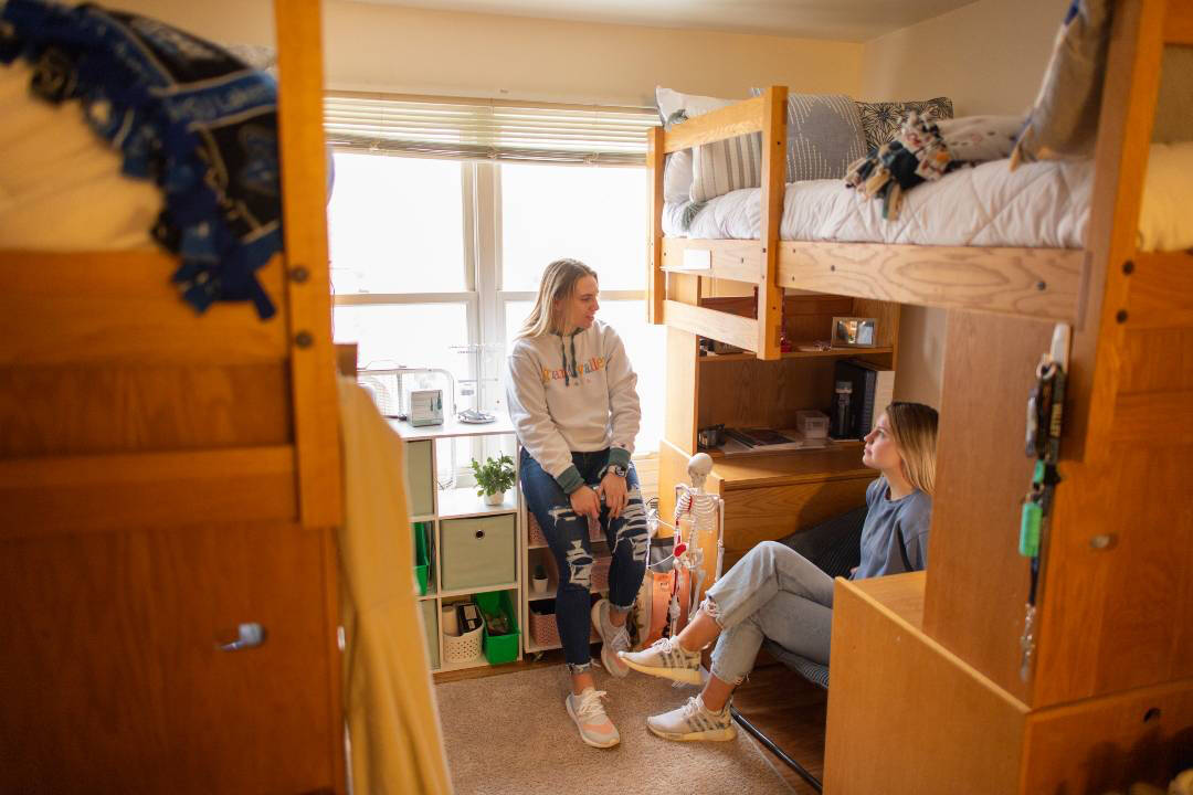 Two students hang out in their bedroom in Kirkpatrick and talk. Two lofted beds, a desk, and storage are in the background.